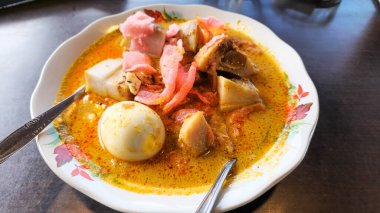Geleneksel bir yemek olan Endonezya Lontong Sayur 'unun yakın plan fotoğrafı. Tabakta uzun pirinç kekleri, haşlanmış yumurta, tavuk ve çıtır krakerler var. Hepsi zengin, lezzetli bir köri sosuna batırılmış. Tabak beyaz bir tabakta sunulur.
