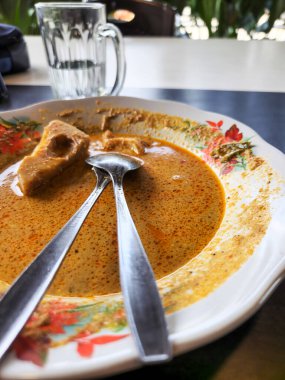 Körili, kremalı, aromalı bir sos ve bir parça yiyecek içeren yakın çekim bir tabak et ya da tofu. Tabakta iki kaşık dinleniyor. Arka planda bir bardak su duruyor. Plakanın çiçek deseni var..