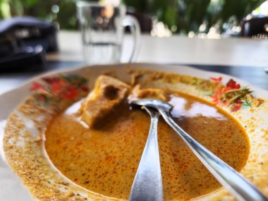 Bir tabak körili soya peyniri ve körinin içinde dinlenen iki kaşık. Plakanın çiçekli bir tasarımı var ve az kullanılmış. Köri zengin, turuncu bir renktir ve kalın bir dokusu vardır. Arka planda bir bardak su var..