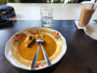 Tepeden çekilen bir fotoğraf, Endonezya köri tabağını koyu ahşap bir masada çiçek desenli bir tabakta yakalar. Yemeğin üzerine iki kaşık konmuştu. Tabağın yanında temiz bir bardak su ve uzun bir bardak buzlu kahve var.. 