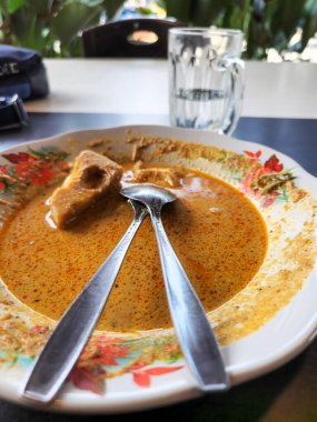 İçinde bir parça tofu olan Endonezya köri tabağının yakın plan çekimi. Tabakta iki kaşık dinleniyor. Arka planda berrak bir bardak su duruyor. Plakası beyaz, çiçek desenli..