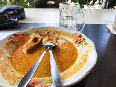 Ortasında bir parça tofu olan bir tabak Endonezya köri tabağı. Yemek kalın ve lezzetli bir köri sosuyla çevrili. İki kaşık tabakta dinleniyor. Tabağın yanında bir bardak su var. Masa koyu renkli..