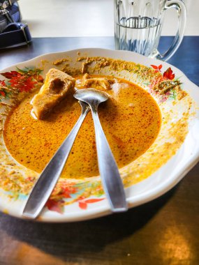 Bir tabak pirinç keki ve iki kaşıkla bir tabak Endonezya köri sergilenen bir fotoğraf. Tabak zengin, turuncu renkli köri sosuyla yıkanır. Yakınlarda bir bardak su var, yemek takımını işaret ediyor. Plakada çiçek tasarımı var..