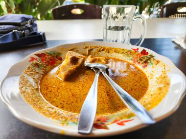 Endonezya körisinin olduğu bir tabağın yukarıdan çekimi. Plakanın çiçekli bir tasarımı var. Tabakta iki kaşık var. Arka planda bir bardak su ve mavi bir torba parçası var. Yiyecekler çoğunlukla yenir..