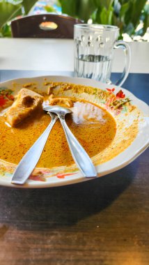 İki çapraz kaşık ve bir bardak suyla birlikte çiçek desenli bir tabakta servis edilen geleneksel Endonezya köri yemeğinin yakın çekimi. Köri, görünen baharatlarla zengin, altın renginde ve bir parça et görünür..