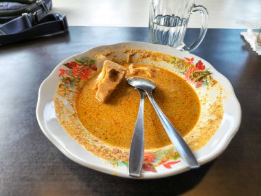 Endonezya Soto yemeğinin iştah açıcı bir tabağı, içinde iki kaşıkla servis edilen bir parça tofu ile zengin, sarı bir çorba var. Yemek beyaz bir tabakta sunuluyor. Renkli bir çiçek deseniyle süslenmiş..