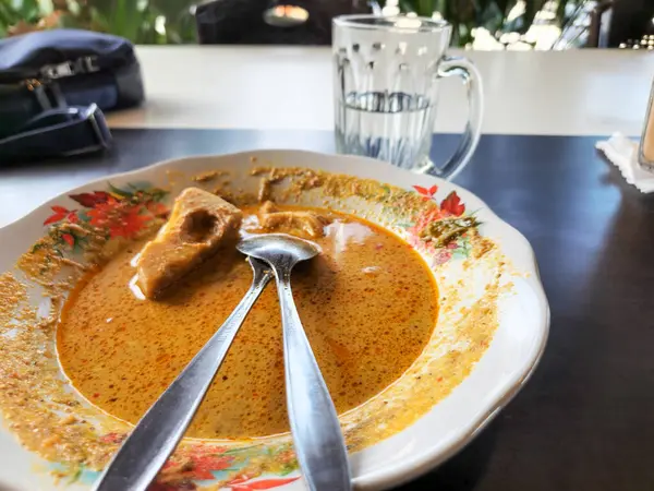 Ortasında bir parça tofu olan bir tabak Endonezya köri tabağı. Yemek kalın ve lezzetli bir köri sosuyla çevrili. İki kaşık tabakta dinleniyor. Tabağın yanında bir bardak su var. Masa koyu renkli..