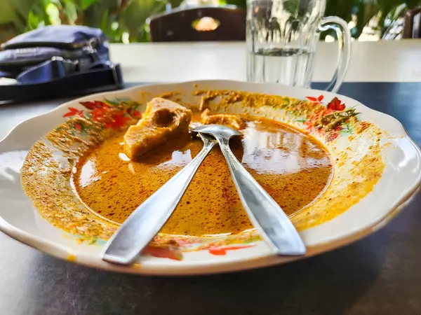 İçinde bir parça et ve aromalı portakal sarısı çorba bulunan köri tabağı ile yakın plan bir fotoğraf. Tabakta iki kaşık dinleniyor. Plakanın kenarlarında çiçek desenleri var. Arka planda bir bardak su ve bir çanta var..