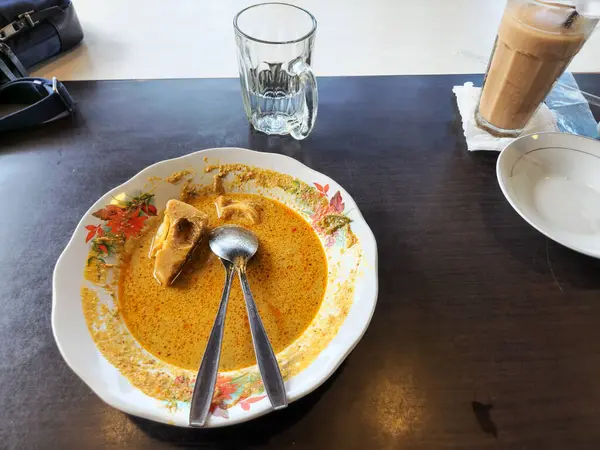 Bir tabak Endonezya yemeği, bir bardak buzlu kahve ve koyu bir masada bir bardak su. Tabakta kalın, sarı soslu bir tabak ve bir parça tofu ya da tempeh var. Tabakta iki kaşık var..