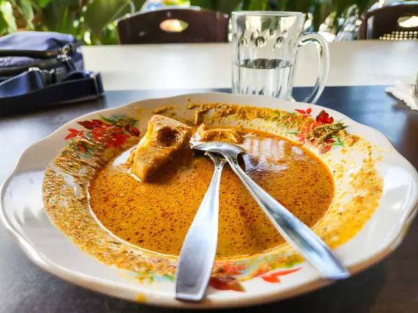 Endonezya körisinin olduğu bir tabağın yukarıdan çekimi. Plakanın çiçekli bir tasarımı var. Tabakta iki kaşık var. Arka planda bir bardak su ve mavi bir torba parçası var. Yiyecekler çoğunlukla yenir..