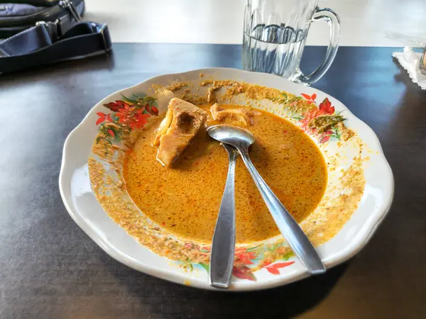 Endonezya Soto yemeğinin iştah açıcı bir tabağı, içinde iki kaşıkla servis edilen bir parça tofu ile zengin, sarı bir çorba var. Yemek beyaz bir tabakta sunuluyor. Renkli bir çiçek deseniyle süslenmiş..