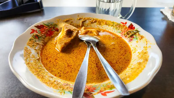 Tepeden çekilen bir fotoğraf Endonezya köri tabağını yakalıyor, kalan tofu parçalarını ve aromalı sosu sergiliyor. Tabağın içinde iki kaşık dinleniyor, bu da yemeğin bittiğini gösteriyor. Tabağın kenarlarında köri kalıntıları var.