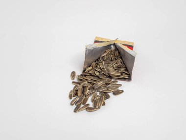 Yakın plan çekimde bir paket ayçiçeği çekirdeği gösterilir. Tohumlar sade beyaz bir zemine saçılır. Paket kısmen açık, içerideki tohumların bolluğunu gösteriyor..