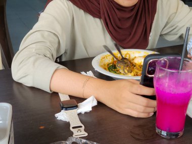 Bir kişi bir masada oturur, bir yemeğe katılır. Bir tabak yiyecekleri, bir bardak canlı pembe içecekleri ve pipetleri var ve ellerinde akıllı bir telefon var. Masada bir elma saati dinleniyor..