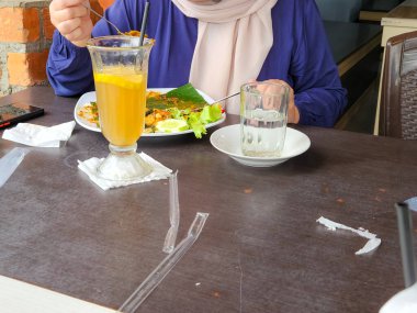 Bir kişi restoranda bir masada oturmuş muz yaprağıyla servis edilen bir tabakta yemek yiyor. Masada iki içki var, biri limon dilimi ve pipetle uzun bir bardakta, diğeri de su dolu basit bir bardakta..