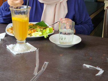 Bir insan bir masada oturmuş, yemeğin tadını çıkarıyor. Bir bardak su ve limon dilimli bir bardak sarı içecek var. Yeşillikli bir tabak yemek ve bir çeşit yemek gibi görünen bir şey, belki de deniz ürünleri..