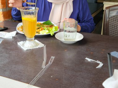 Bir insan açık hava yemek ortamında bir masada oturur. Masada bir bardak limon dilimleri, bir bardak su ve bir tabak yiyecek yer alıyor. Masada pipetler ve peçeteler de var. Bu da sıradan bir yemek olduğunu gösteriyor..