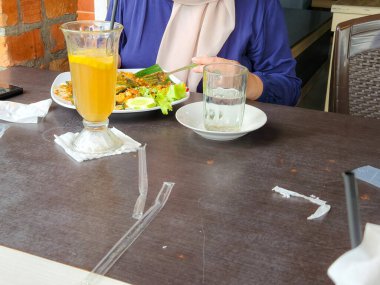 Yemek sahnesinde tesettürlü bir insan bir restoranda yemeğin tadını çıkarıyor. Masada bir tabak yiyecek, bir bardak meyve suyu ve bir bardak su var. Masada kırık plastik pipetler görülüyor. Bu da yeni bir içki olduğunu gösteriyor..