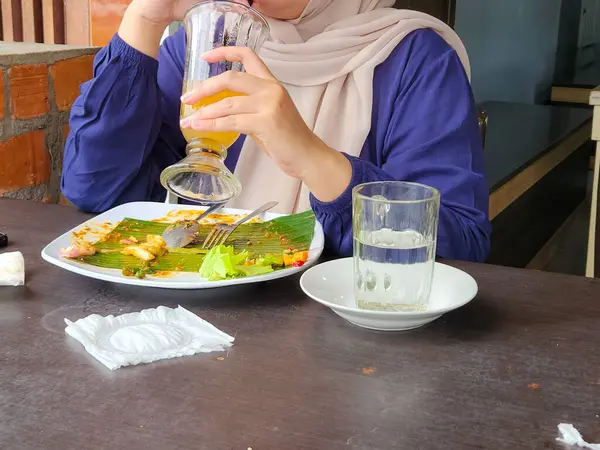 Bir kadın yemeğini bitirdikten sonra bir restoranda içki içerken görülüyor. Görüntü bir rahatlama ve tatmin anı yakalar. Muz yaprağında kısmen yenmiş bir tabak, bir bardak su ve bir peçete masanın üzerinde..