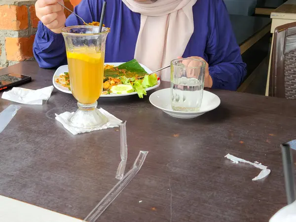 Bir kişi restoranda bir masada oturmuş muz yaprağıyla servis edilen bir tabakta yemek yiyor. Masada iki içki var, biri limon dilimi ve pipetle uzun bir bardakta, diğeri de su dolu basit bir bardakta..