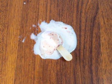 Tepeden çekimde, erimiş bir dondurmanın ahşap bir yüzeyde yattığını görüyoruz. Buzlu çubuk kısmen erimiş ve kalıntıları ahşap çubuğun etrafında bir su birikintisi oluşturmuş. Yüzey, zıt bir zıt zemin oluşturan ılık tonlu ahşap bir tahıldır.. 