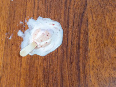 Kahverengi ahşap bir yüzeyde duran erimiş dondurmanın yukarıdan aşağı görüntüsü. Buzlu dondurma kısmen erimiş, geriye yapışkan, beyaz ve pembe bir su birikintisi bırakmış. Ahşap yüzeyin doğal bir ahşap deseni vardır. Bu da sahnenin dokusuna zıtlık katar.. 