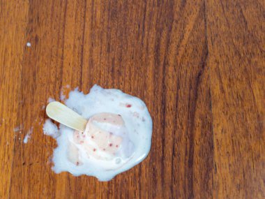 Yakın çekim, kahverengi ahşap bir yüzeye dökülen dondurmayı yakalar. Erimiş dondurma, görünen kırmızı meyve parçalarıyla buzlu çubuğun etrafına yayılmış. Görüntü bir karmaşa hissi veriyor, yaz ve kaçırılan bir ikram..