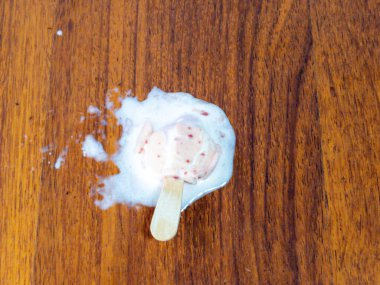 Tepeden aşağı bir çekim, ahşap bir yüzey üzerinde erimiş dondurmayı gösteriyor. Buzlu şeker kısmen erimiş ve etrafında su birikintisi oluşturmuş. Tahta tohumu sıcak bir zemin sağlıyor.