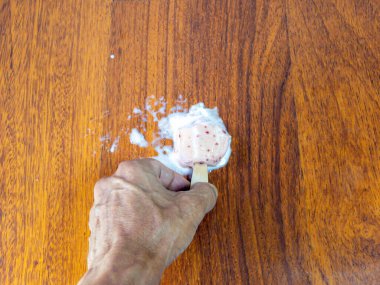 Yakın çekim, kısmen erimiş bir dondurma çubuğunu tutan bir eli gösteriyor. Ahşap bir masada kırmızı benekler var. Dondurma damladı ve yayıldı, bu da ortalığı karıştırdı..