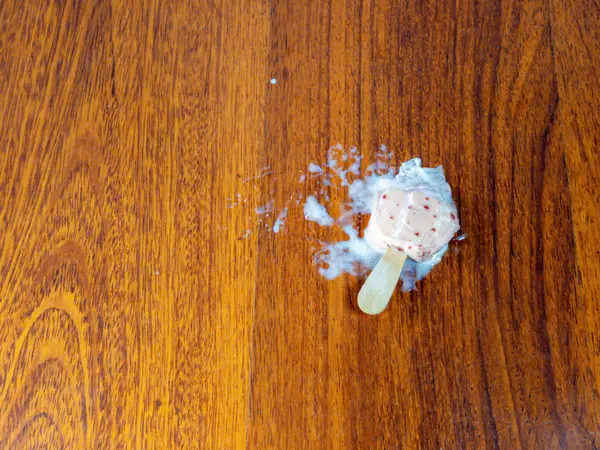 Tepeden çekilen bir çekim, parlak ahşap bir yüzeyde eriyen buzlu dondurmayı yakalar. Dondurma şeklini kaybetmiş ve erimiş krema birikintisiyle çevrili. Tahta dokular, yaz ziyafeti kazasının altını çizerek sıcak bir zemin sağlıyor..