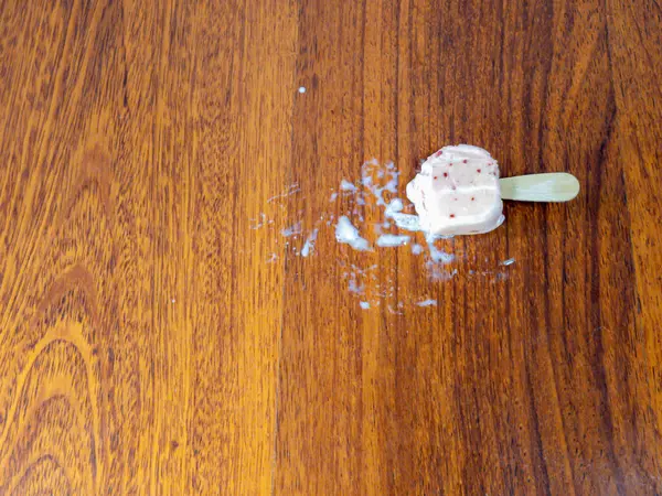 Yakın çekim, kısmen erimiş çilekli dondurmayı parlak ahşap bir yüzeyde yatarken yakalar. Dondurma eridi, bir su birikintisi ve ahşap üzerinde izler bıraktı. Tahta çubuk hala dondurmaya bağlı..