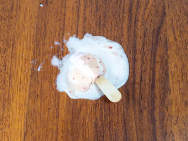 Tepeden çekimde, erimiş bir dondurmanın ahşap bir yüzeyde yattığını görüyoruz. Buzlu çubuk kısmen erimiş ve kalıntıları ahşap çubuğun etrafında bir su birikintisi oluşturmuş. Yüzey, zıt bir zıt zemin oluşturan ılık tonlu ahşap bir tahıldır.. 