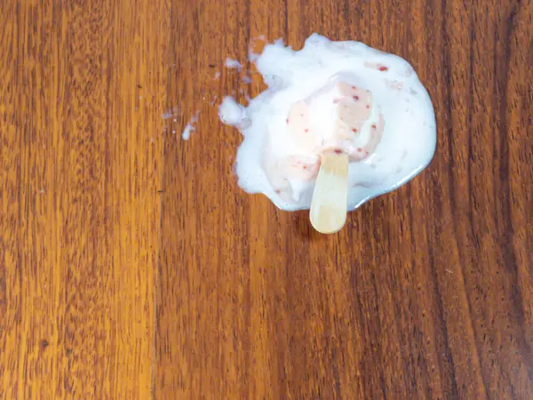 Tahta bir yüzeyde erimiş buzlu şeker sıcak bir yaz gününü işaret ediyor. Görüntü buzlu dondurmanın kalıntılarını ve erimiş dondurmayı gösteriyor. Ahşap arka plan sahneye doğal ve sıcak bir ton ekler..