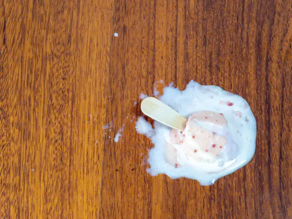 Tepeden aşağı bir atış, yere dökülen dondurmayı ahşap bir yüzeyde yakalar. Tahta çubuklu eritilmiş dondurma su birikintisinde yatıyor. Görüntü, ahşap ve dondurmanın karışıklığına ve dokusuna odaklanıyor..