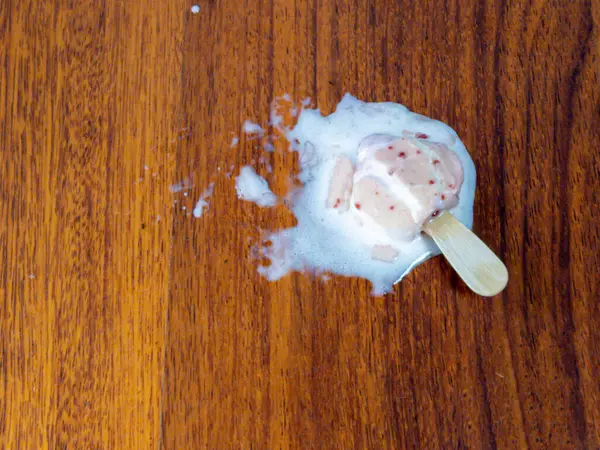 Yakın çekim, erimiş dondurmayı yakalar ve sıcak tonlu ahşap bir yüzeyde yatar. Dondurma tamamen erimiş ve tahta bir sopa hala üzerinde. Görüntü, yaz mevsimi, sıcaklık ve geçici bir zevk hissi veriyor.. 