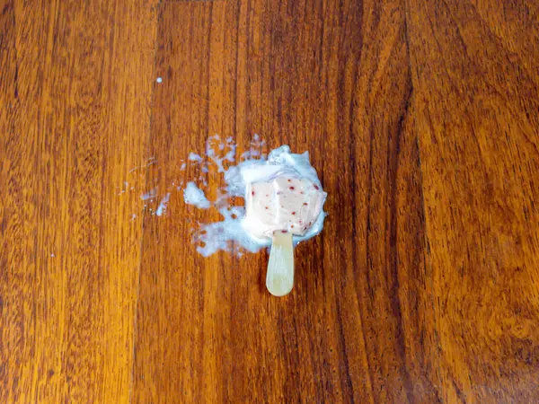 Yukardan aşağı bir atış, erimiş dondurmayı sıcak ahşap bir yüzeyde yakalar. Dondurma, görünen kırmızı benekleri ile, tamamen erimiş, odunlara yayılmış. Görüntü, yaz sıcağı ve geçici bir zevk hissi veriyor..