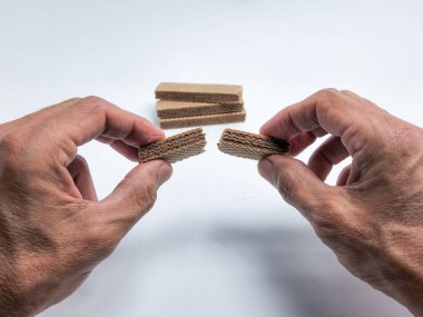 Gofretli kurabiyeyi ikiye bölen ellerin yakın çekimi. Görüntü, çerezin dokusunu ve onu kırma eylemini vurgulayan temiz beyaz bir arkaplana göre ayarlanır. Ek gofretler arka planda istiflenmiş.