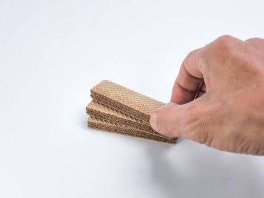 Bir elin çikolatalı gofret kurabiyelerini düz beyaz bir zemine tutarken yakın plan çekimi. Gofretler dikdörtgendir, waffle gibi bir dokusu ve çikolata dolu katmanları vardır. Resim, kurabiyelerin dokusunu ve rengini vurgular.