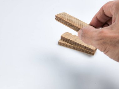 Bir el çikolatalı gofret kurabiyesini bir yığın benzer kurabiyenin üzerinde tutuyor, hepsi de temiz beyaz arka plana karşı. Görüntü, onların cazibesini vurgulayarak, gofretli kurabiyelerin dokusunu ve katmanlarını vurguluyor. Kompozisyon basit..