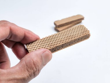 Yakın çekim, dikdörtgen gofretli kurabiye tutan bir eli yakalar. Kurabiye kahverengi, katmanlı bir dokusu var. Arka planda iki ekstra kurabiye bulanıklaşıyor. Arka plan düz beyaz, temiz ve düzensiz bir estetik sağlıyor..