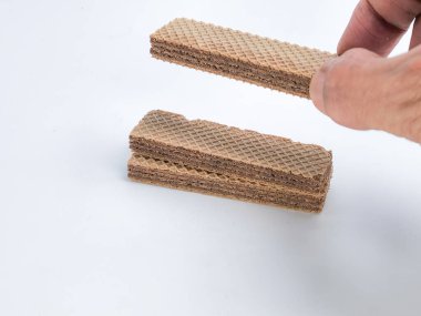 Yakından çekilen bir fotoğrafta, beyaz bir yüzeyde iki yığın gofret bulunan bir çikolatalı gofret tutan bir el görülüyor. Gofretlerin çikolata dolgulu katmanlı bir dokusu var. Görüntü iyi ışıklandırılmış, beyaz bir arkaplanı var ve gofretleri vurguluyor..