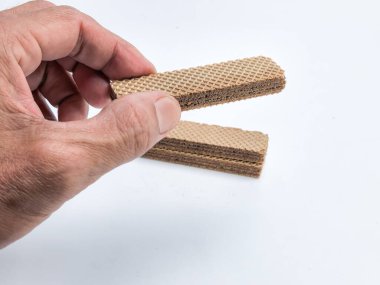 Yakından çekilen bir fotoğraf çikolatalı gofret tutan bir el ve altında da bir yığın gofret var. Görüntü temiz beyaz bir arkaplan üzerinde yakalanıyor, gofretlerin dokusu ve rengi vurgulanıyor.