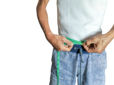 Yeşil bir mezura ile belini ölçen bir adamın yakın plan fotoğrafı. Beyaz bir tişört ve mavi desenli şort giyiyor. Görüntü, sağlık, kilo kaybı ve vücut imajının ölçülmesi, vurgulanması hareketini vurgular..