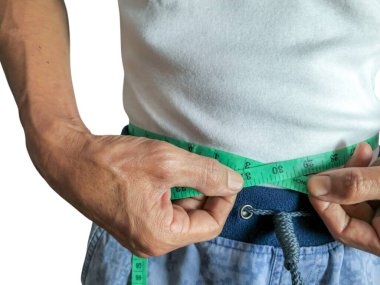 Yeşil bir mezura ile belini ölçen bir adamın yakın plan çekimi. O, beyaz bir tişört ve mavi pantolon giyiyor. Görüntü, ağırlık yönetimi, sağlık ve vücut büyüklüğü kavramını vurgulayarak erkeğin ellerine ve mezuraya odaklanıyor..