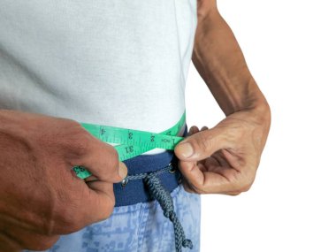 Yeşil bir mezura ile belini ölçen bir adamın yakın plan çekimi. O, beyaz bir atlet ve mavi askılı pantolon giyiyor. Görüntü, muhtemelen kilo verme hedefleri veya vücut sağlığı izleme faaliyetlerini gösteren ölçüm sürecine odaklanır. 