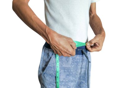 Yaşlı bir adam bel ölçüsünü yeşil bir mezura ile ölçer, sağlık ve sağlığa odaklandığını gösterir. Beyaz bir tişört ve mavi desenli pantolon giyiyor. Resim vücut ölçümlerini izleme eylemini vurgular.