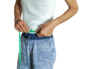 Yaşlı bir adam yeşil bir mezura ile belini ölçer. Beyaz bir tişört ve mavi desenli şort giyiyor. Görüntü, iyi ışıklandırma ve odaklanma ile beyaz bir arkaplanda izole edilmiştir. Adamın elleri ve kolları görünür..