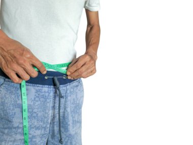 Yaşlı bir adam yeşil bir mezura ile belini ölçüyor. O, beyaz bir tişört ve asma ipli mavi desenli şort giyiyor. Görüntü belden yukarı, beyaz arkaplanlı..