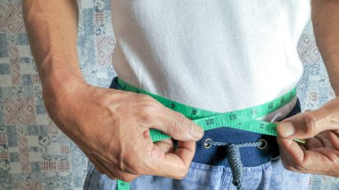 Yeşil bir mezura ile belini ölçen bir adamın yakın çekim görüntüsü. Adam beyaz bir tişört ve mavi bir şort giyiyor. Arka plan desenli bir duvar. Görüntü, görünürdeki ellerle bel ölçümüne odaklanır..