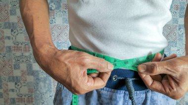 Yakın çekim, yeşil bir mezura ile belini ölçen birini yakalar. Kişi beyaz bir tişört ve mavi bir şort giyiyor. Elleri bandı dikkatlice tutuyor ki doğru ölçebilelim. Odaklanma bel hizasında..